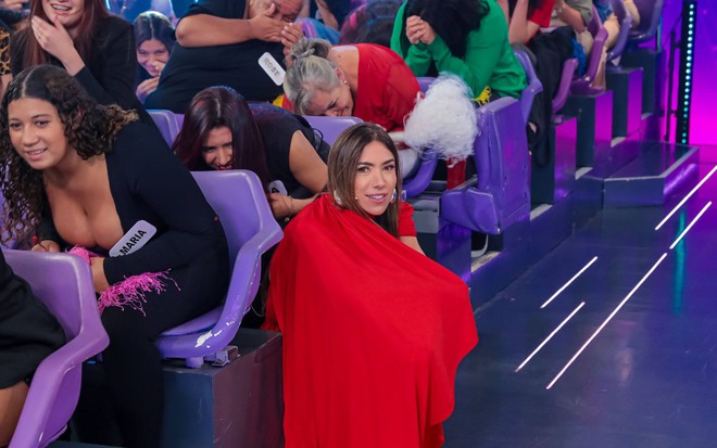 Mulher com microfone preso à roupa vermelha se destaca na plateia de programa; outras pessoas riem e se abaixam nas cadeiras lilás.