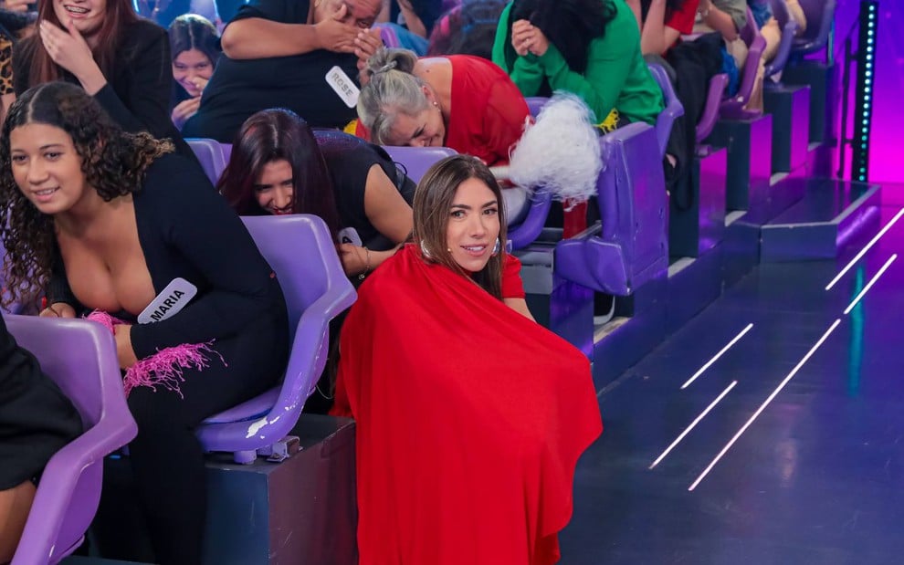 Mulher com microfone preso à roupa vermelha se destaca na plateia de programa; outras pessoas riem e se abaixam nas cadeiras lilás.