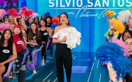 Apresentadora em palco de auditório sorrindo com plateia animada ao fundo, segurando pompons coloridos, em cenário de programa de TV.