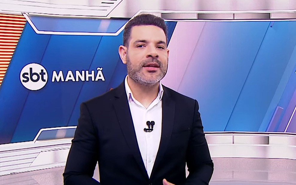 Apresentador sorri em estúdio do SBT Notícias; veste terno azul e camisa branca, com microfone preso na lapela.