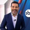 Apresentador sorri em estúdio do SBT Notícias; veste terno azul e camisa branca, com microfone preso na lapela.