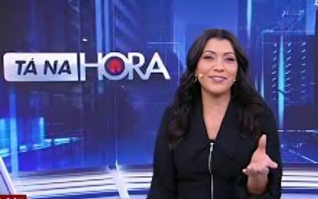 Mulher apresenta programa de TV com cenário moderno e azul ao fundo; ela usa roupa preta e está com microfone de lapela. Ao lado, lê-se “TÁ NA HORA” em destaque.