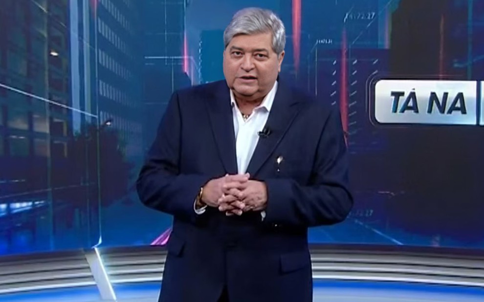 Homem de cabelos grisalhos e terno azul escuro apresenta um programa em um estúdio com fundo digital. Texto parcial: "TÁ NA".