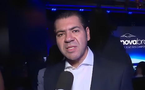 Daniel Abravanel fala em microfone e olha para a câmera com expressão de contragosto