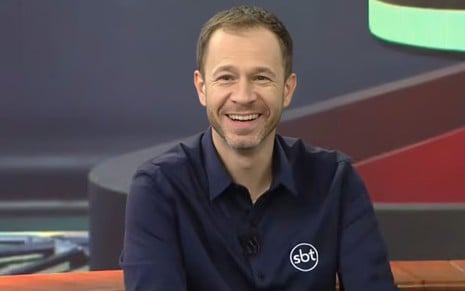 Homem sorridente com barba rala veste camisa azul-marinho com logotipo do SBT; cenário colorido ao fundo