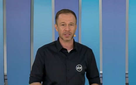 Homem em frente a fundo azul, usando camisa preta com logotipo do SBT. Ele está com microfone de lapela e expressão séria.