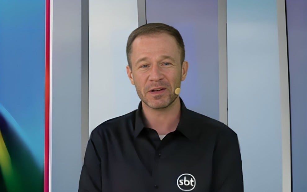 Homem com camisa preta do SBT e microfone de orelha, posicionado em frente a um fundo colorido e geométrico.