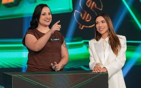 Duas mulheres em um programa de TV: uma à esquerda com camiseta marrom, sorrindo e apontando; outra à direita de terno branco, sorrindo.