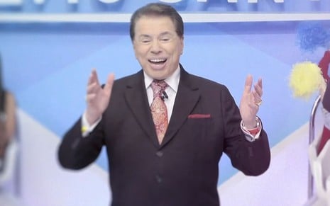 Silvio Santos com as mãos levantadas e sorrindo no cenário do Programa Silvio Santos