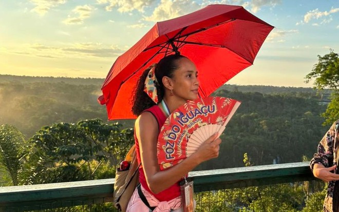 Taís Araujo segura um guarda-chuva e um leque vermelhos; ela está nas Cataratas do Iguaçu, no Paraná