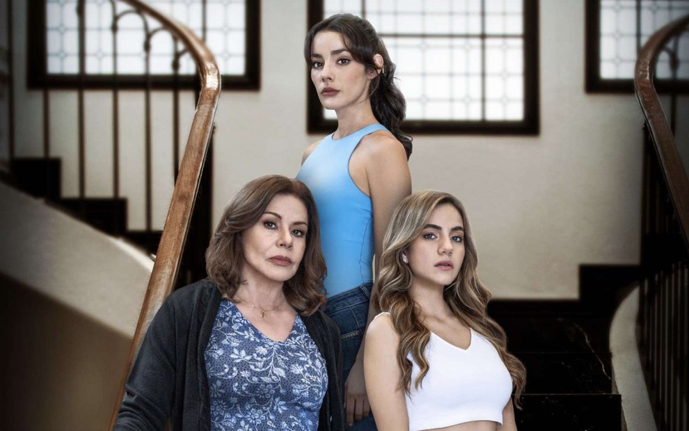 Três mulheres, uma mais velha e duas jovens, posam em uma escadaria com expressões sérias