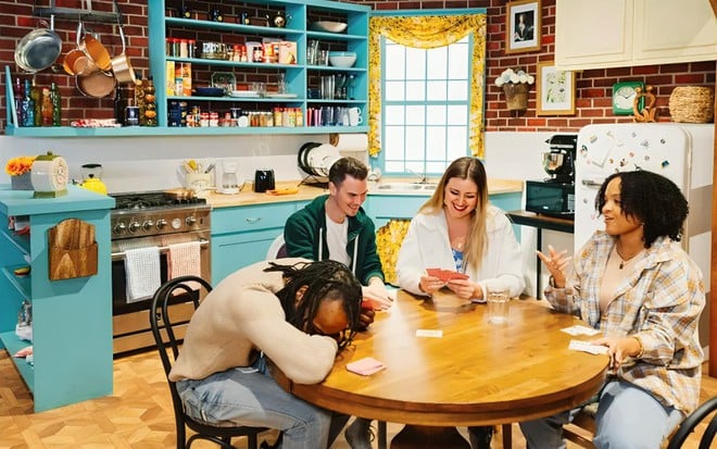 Quatro pessoas estão sentadas à mesa de uma cozinha cenográfica, rindo bastante