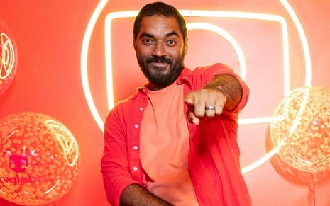 Thomás Aquino todo vestido de laranja, em cenário totalmente laranja, com o logo da Globo em led ao fundo