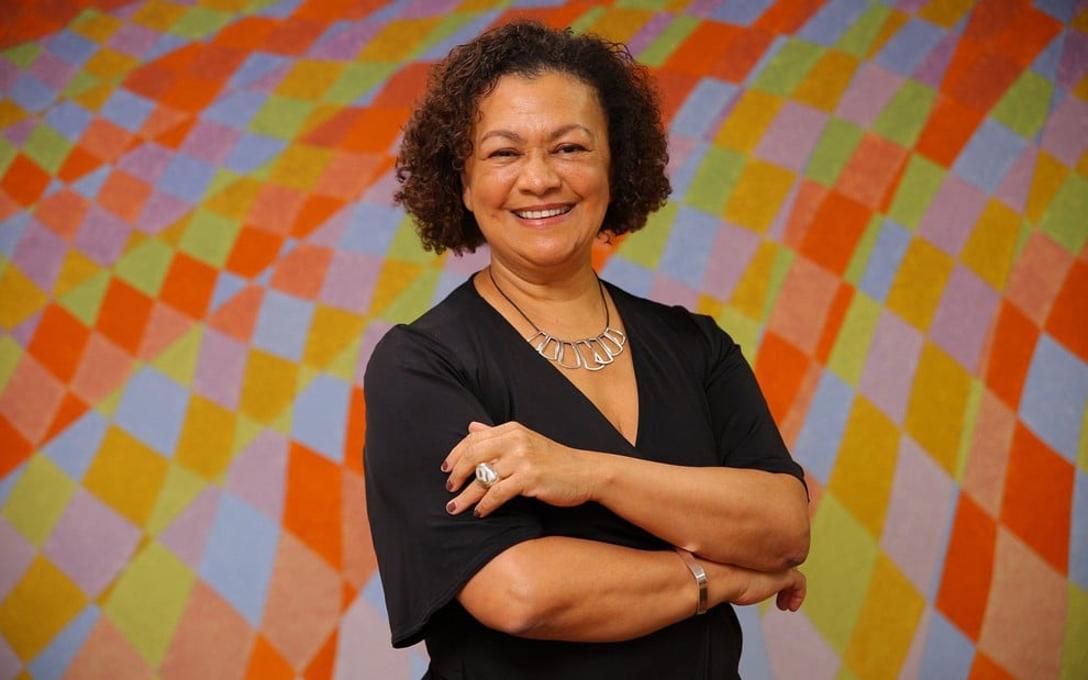 Maria Angela de Jesus posa sorridente, com os braços cruzados, na frente de um fundo colorido