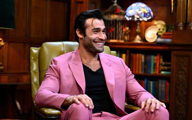 Com terno cor-de-rosa, Sam Asghari sorri abertamente na sala de reuniões do reality The Traitors