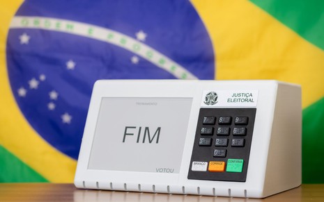 Urna eletrônica da Justiça Eleitoral com a palavra "FIM" exibida na tela, em frente à bandeira do Brasil desfocada ao fundo