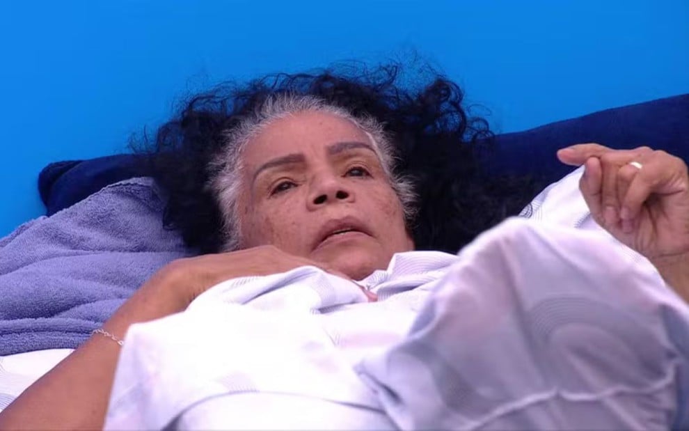 Vilma Nascimento deitada no quarto Fantástico do BBB 25, enterrada embaixo de um edredom branco