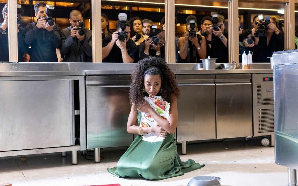 Gabz em cena de Mania de Você em que Viola sofreu derrocada; ela está ao chão, segurando livro de receitas, observada por fotógrafos