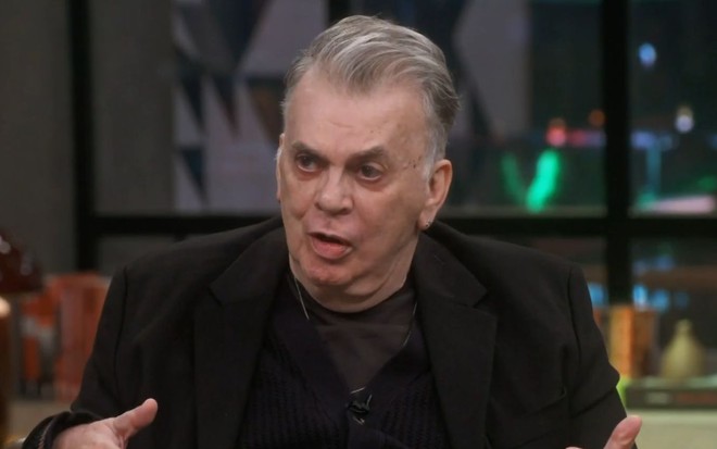 Walcyr Carrasco usando terno preto, sentado no cenário do Conversa com Bial