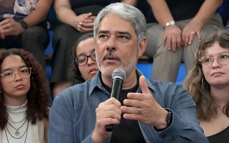 William Bonner no cenário do Altas Horas, com camiseta preta e camisa azul, segurando um microfone