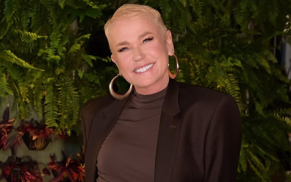 Xuxa Meneghel sorri em frente a painel com plantas; ela usa roupa preta e brinco de argola