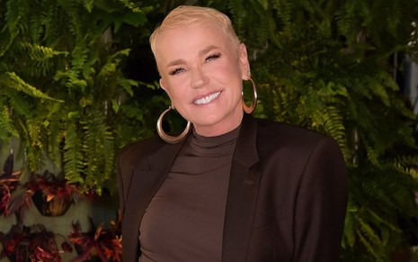 Xuxa Meneghel sorri em frente a painel com plantas; ela usa roupa preta e brinco de argola