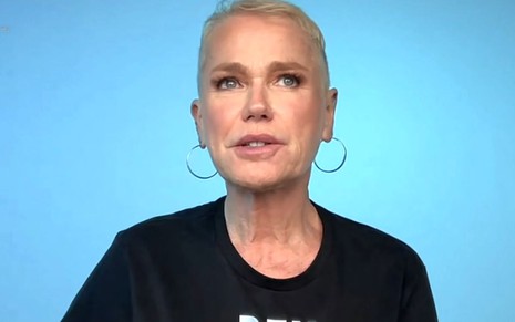 Xuxa Meneghel usa camiseta preta e está em frente a um fundo azul