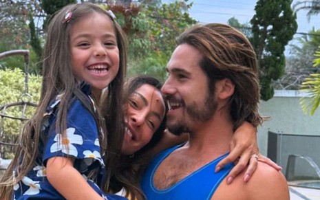 Nicolas Prattes segurando Zoe no colo, enquanto a menina dá um largo sorriso e Sabrina Sato abraça os dois