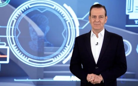 Homem de terno azul fala em estúdio com fundo tecnológico e imagem de corpo humano digitalizado.