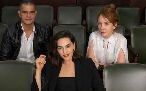 Sentados em uma espécie de plateia de cinema estão os atores Eduardo Moscovis, Tainá Müller e Camila Morgado