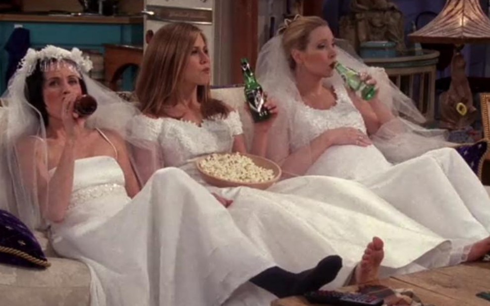As atrizes Courteney Cox, Jennifer Aniston e Lisa Kudrow sentadas no sofá vestidas de noiva e bebendo cerveja em cena de Friends