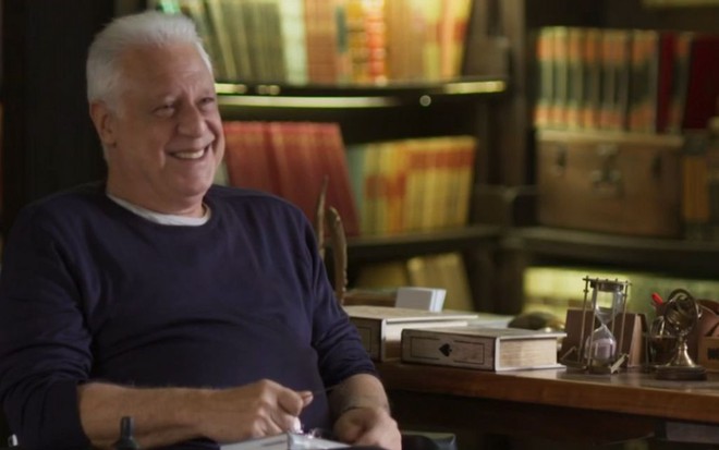 O ator Antonio Fagundes em cena da novela Bom Sucesso como o personagem Alberto, sentado em um cenário de biblioteca