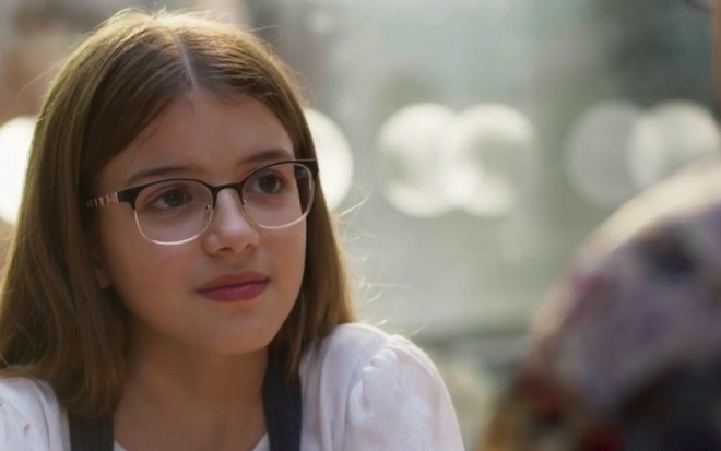 A atriz mirim Valentina Vieira vestindo uma camiseta branca e usando óculos, em cena da novela Bom Sucesso, da Globo