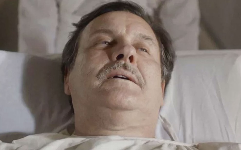 O ator Antonio Calloni caracterizado como o Júlio de Éramos Seis em uma maca de hospital, com rosto lívido e expressão distante, como se tivesse prestes a morrer