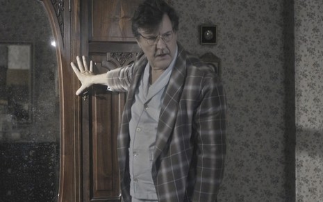 O ator Antonio Calloni de pijamas caracterizado como o Júlio em cena de Éramos Seis, ele se apoia em um guarda-roupa, visivelmente debilitado
