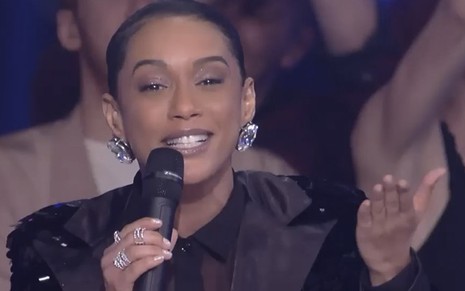 A atriz e apresentadora Taís Araujo em um look preto brilhante, segura um microfone no palco do Popstar