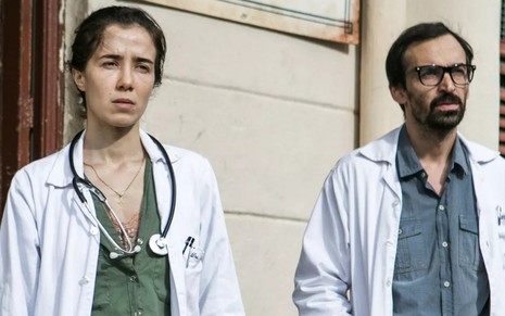 Com avental de médico, Carolina (Marjorie Estiano) e Evandro (Julio Andrade) olham para o horizonte do lado de fora de hospital