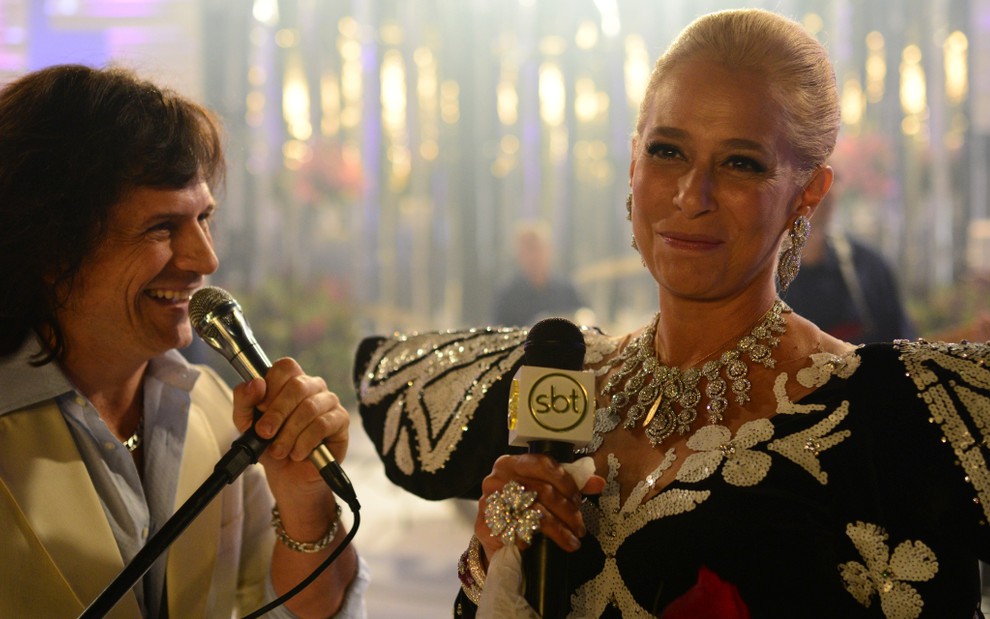 Felipe Rocha (Roberto Carlos) e Andréa Beltrão (Hebe Camargo) em cena do filme Hebe | Ela segura microfone do SBT