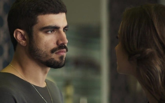 Caio Castro caracterizado como Rock de A Dona do Pedaço, de perfil, olhando para Nathalia Dill, de costas, na pele de Fabiana