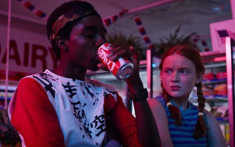 O ator Caleb McLaughlin, observado por Sadie Sink, toma um gole de Coca-Cola em Stranger Things - Reprodução/Netflix