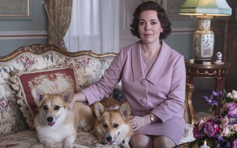 A atriz Olivia Colman sentada no sofá com dois cachorros na terceira temporada de The Crown