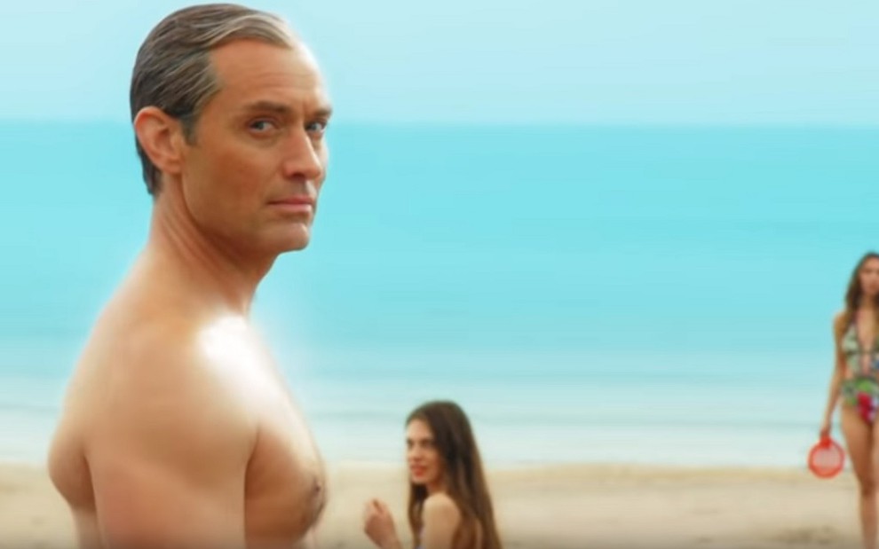 O ator Jude Law em imagem da minissérie The New Pope, sem camisa em uma praia e observado por mulheres à sua volta