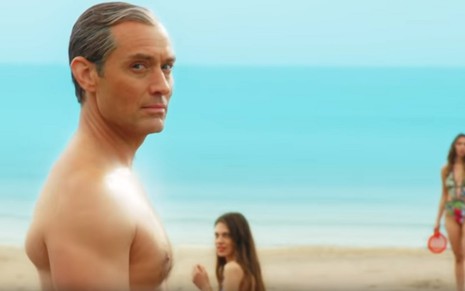 O ator Jude Law em imagem da minissérie The New Pope, sem camisa em uma praia e observado por mulheres à sua volta