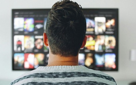 Espectador de costas diante de uma televisão que exibe várias opções de filmes e séries para assistir