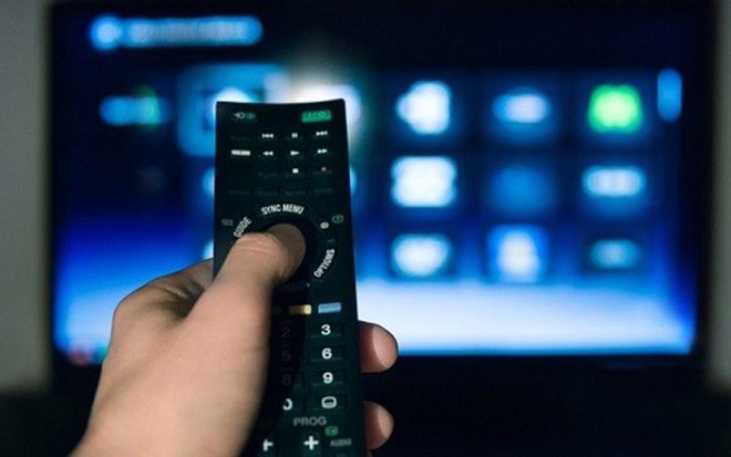 Uma mãe segura um controle em primeiro plano; ao fundo, uma televisão ligada