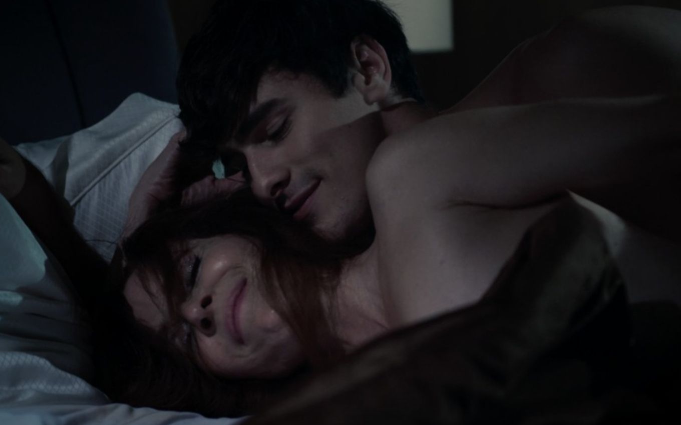 A atriz Deborah Evelyn como Betty está deitada na cama, com um sorriso, enquanto é abraçada por Bruno Montaleone, o Mateus, em cena de Verdades Secretas 2