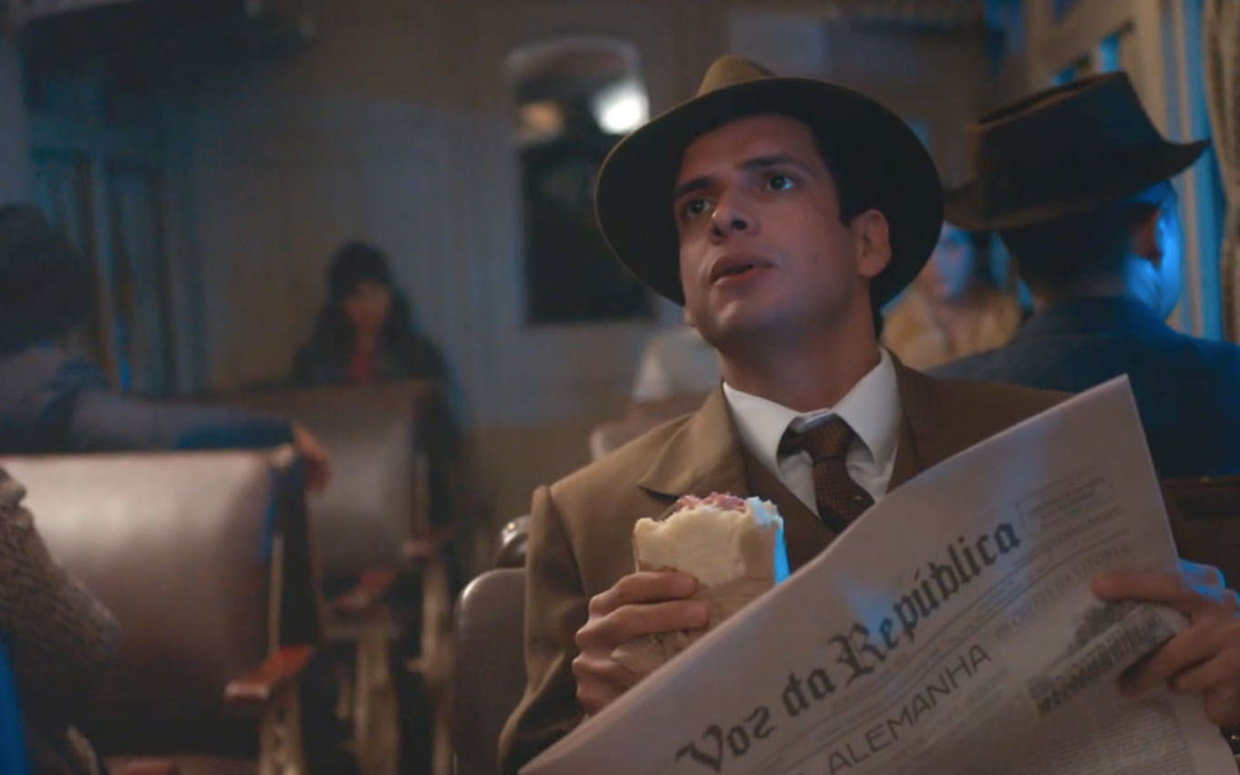 O ator Fabrício Belsoff segura um sanduíche com a mão esquerda e um jornal com a direita em um vagão de trem como o Rafael em cena de Além da Ilusão