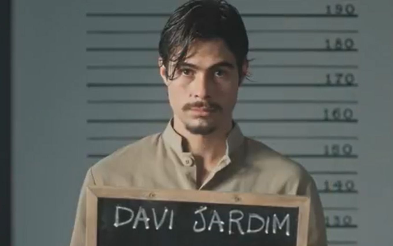 O ator Rafael Vitti como Davi, com uniforme de prisioneiro, segura uma placa de identificação na cadeia em cena de Além da Ilusão