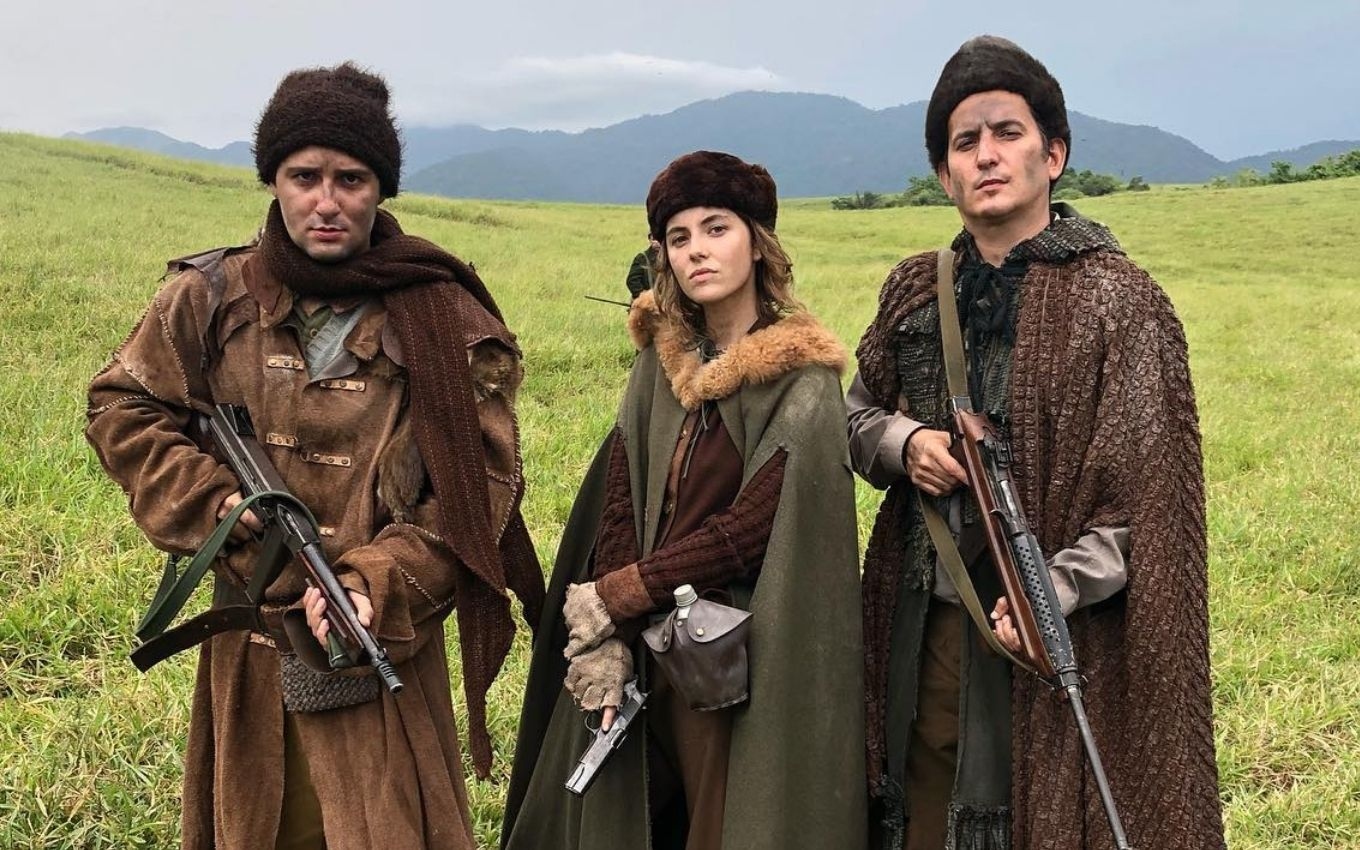 Silvana (Thayla Luz) veste casaco grosso de guerra e segura pistola em cena de Além da Ilusão; há um soldado em cada lado da personagem em cena de Além da Ilusão