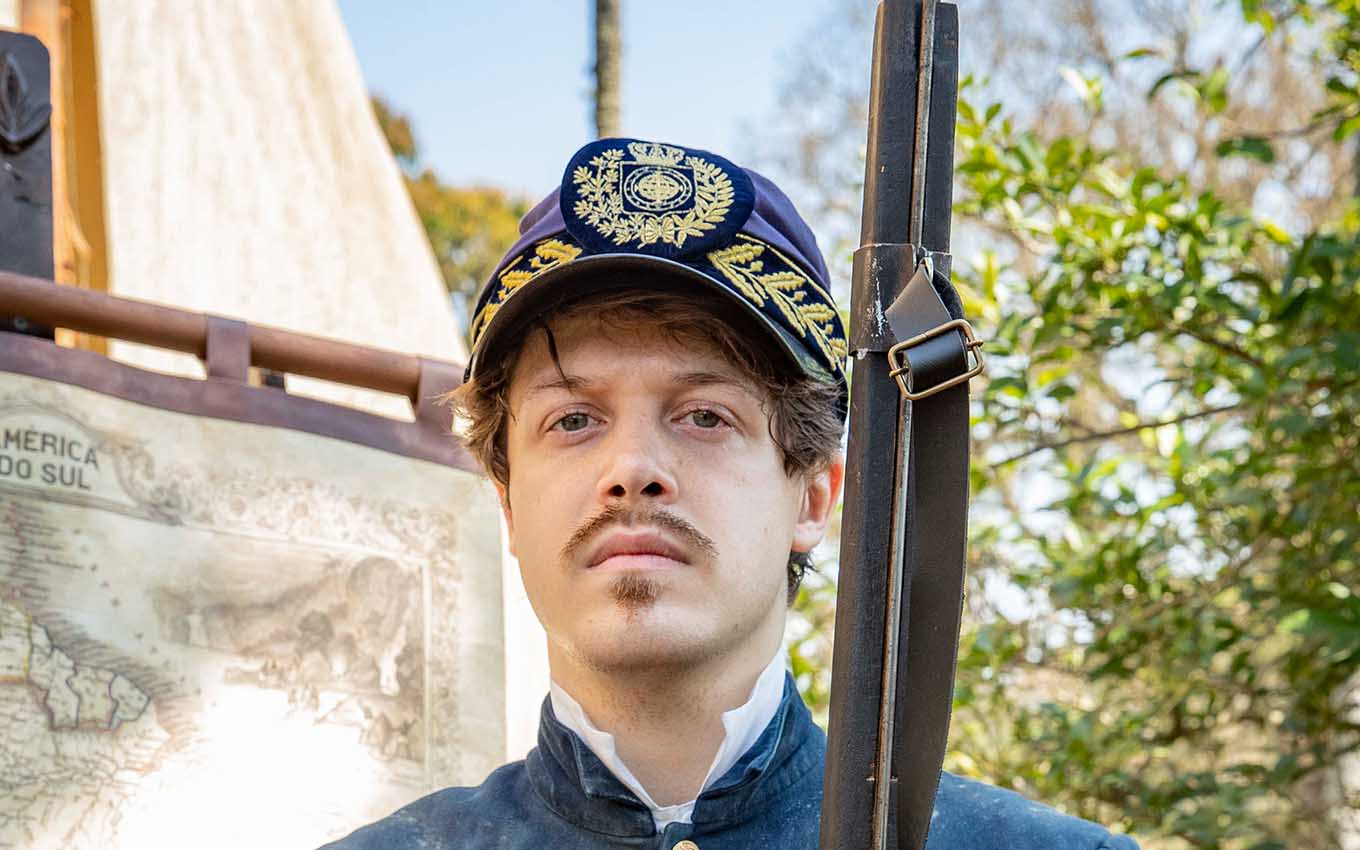 O ator Daniel Torres caracterizado como Gastão com um uniforme militar e segura uma escopeta em Nos Tempos do Imperador